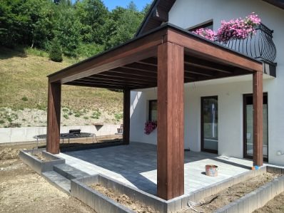 Ciemnobrązowa pergola z płaskim dachem, przylegająca do białego domu z balkonem. Szara kostka brukowa na tarasie, zielone wzgórze w tle.