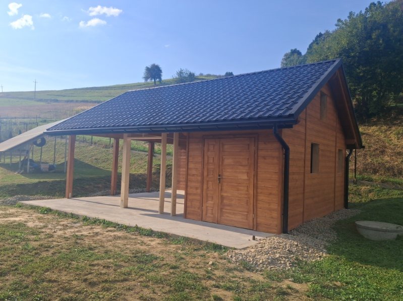 Drewniany budynek z zadaszeniem na betonowej wylewce, otoczony zielenią. Ciemny dach, brązowe drewno, słoneczny dzień. Widok na wzgórza w tle.
