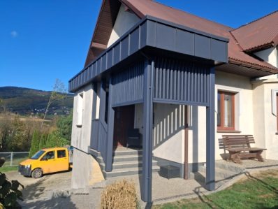 Zadaszone wejście do domu z szarymi, pionowymi panelami i schodami. W tle dom z brązowym dachem, żółty samochód i zieleń. Słoneczny dzień.