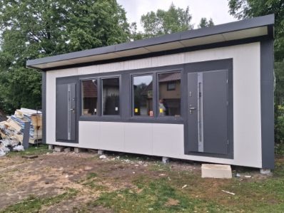 Nowoczesny, parterowy budynek z płaskim dachem, szare drzwi i okna, białe panele ścienne. Widoczne materiały budowlane obok. Budynek na fundamencie.