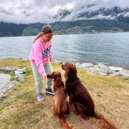 Lena Kowalska - Dziewczyna karmiąca smakołykiem dwa rude irlandzkie setery na tle jeziora i gór w Tychach. Psy na smyczy, jeden siedzi, drugi czeka. Pochmurne niebo.