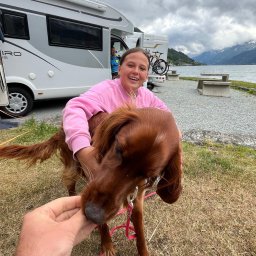 Lena Kowalska - Uśmiechnięta dziewczyna w różowej bluzie przytula rudego setera irlandzkiego, karmionego z ręki na tle jeziora i kampera.