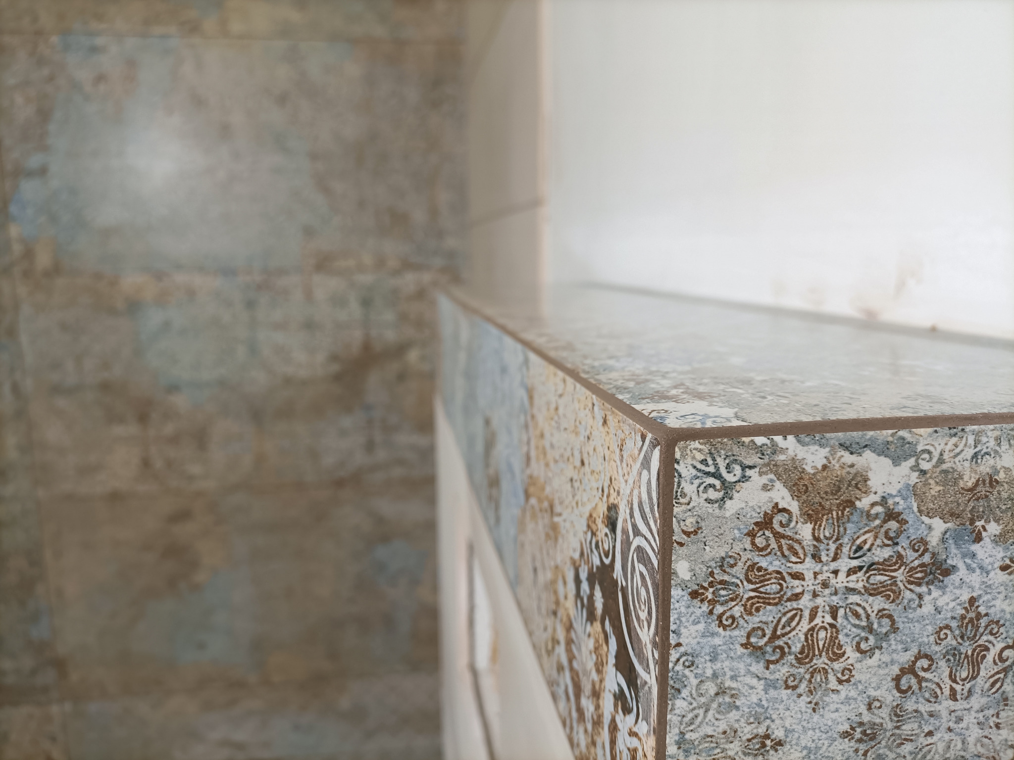 Wykończony fragment łazienki z blatem i ścianą pokrytymi płytkami w odcieniach beżu, błękitu i brązu, z wyraźnym wzorem ornamentacyjnym na krawędzi blatu.