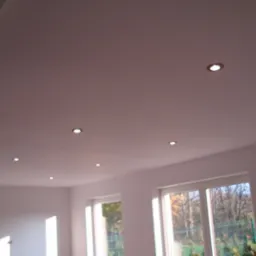 Gładka, pomalowana na jasny odcień sufitu z zamontowanymi punktowymi lampami LED w nowym pomieszczeniu z oknami.