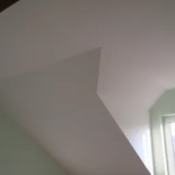 Poddasze po remoncie z ukośnym sufitem, pomalowane na biało, z dwoma wbudowanymi punktowymi lampami LED, ściany w odcieniu jasnej zieleni, okno z widokiem na drzewa bez liści.