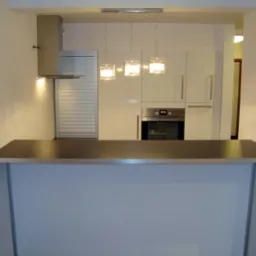 Nowoczesna kuchnia z białymi szafkami, szarym blatem i trzema wiszącymi lampami o kwadratowych abażurach. Widoczny okap i zabudowany piekarnik.