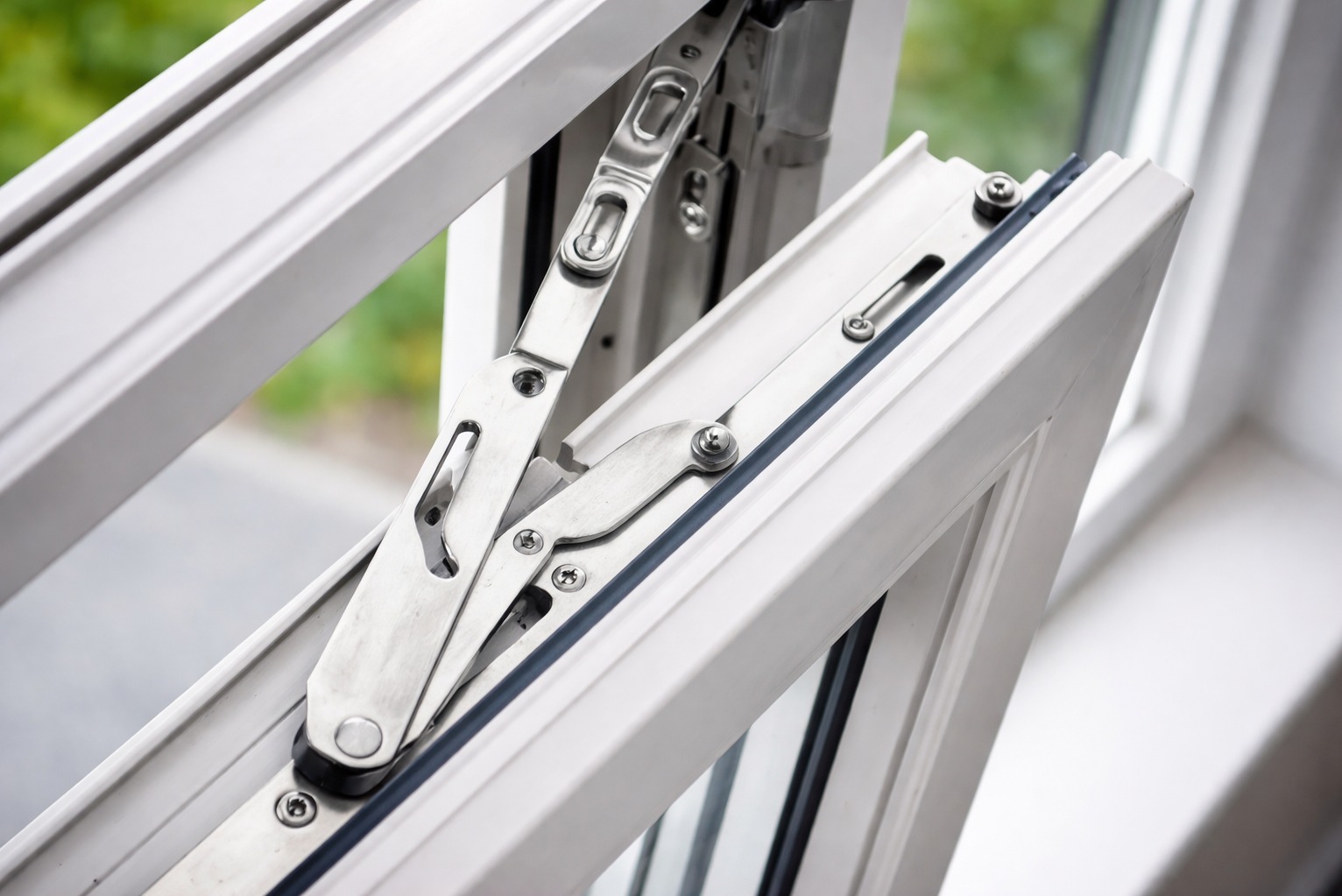 Detaliczne ujęcie mechanizmu okna uchylnego, z widocznymi metalowymi elementami i uszczelką, na tle rozmytego krajobrazu za oknem. Biała rama okienna.