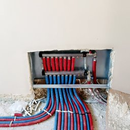 Przemek Szczypiorski - Rozdzielacz ogrzewania podłogowego z rurami w kolorze czerwonym i niebieskim, zamontowany w ścianie. Widoczne elementy instalacji i surowy stan podłoża.