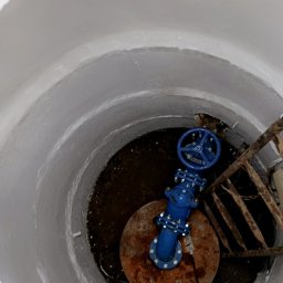 Przemek Szczypiorski - Niebieski zawór w studni z metalową drabinką, widok z góry. Białe, betonowe ściany studni kontrastują z rdzawą pokrywą i ciemnym dnem. Ujęcie z góry.