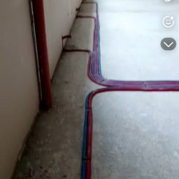 Przemek Szczypiorski - Ułożone rury instalacji hydraulicznej (czerwone i niebieskie) na podłodze w surowym pomieszczeniu. Widoczna drabina w tle.