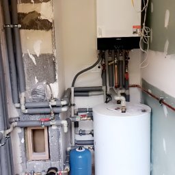 Przemek Szczypiorski - Biały bojler i piec Viessmann z szarymi rurami w pomieszczeniu w trakcie remontu. Widoczne niedokończone ściany i podłoga z jasnych paneli.