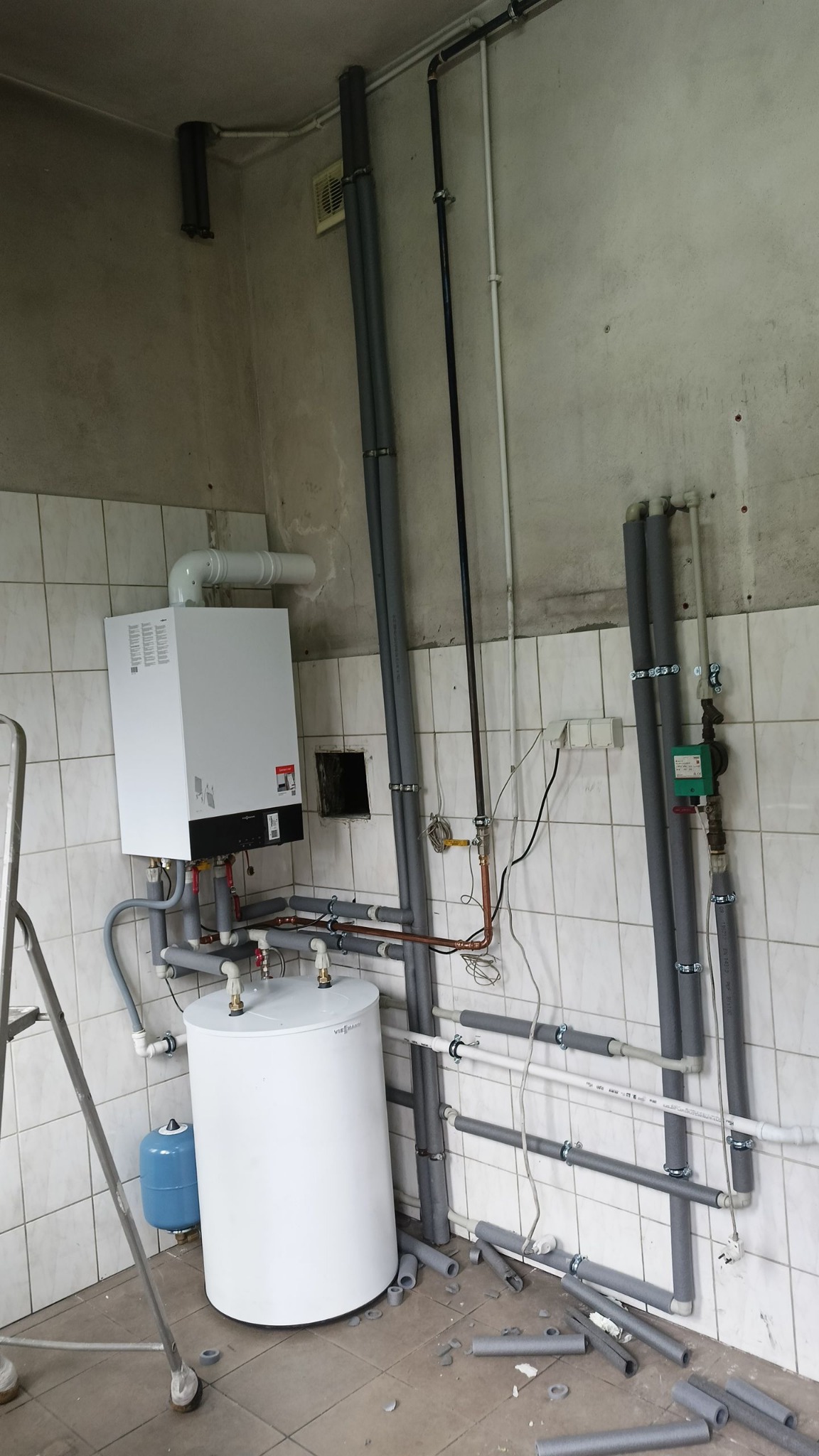 Biały piec i zasobnik ciepłej wody Viessmann z szarymi rurami w izolacji, na tle białych płytek. Widoczne elementy instalacji hydraulicznej i grzewczej w kotłowni.
