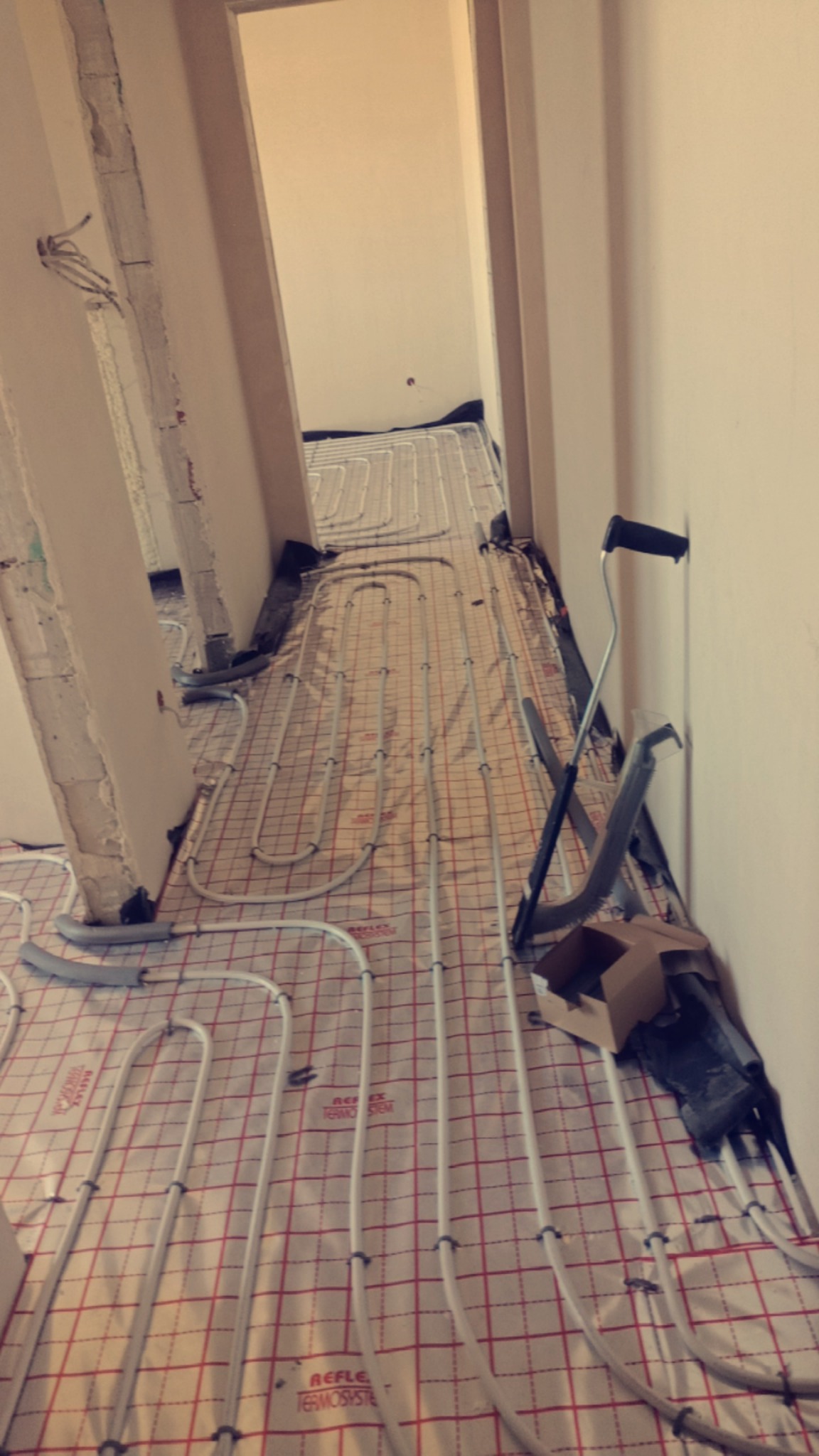 Białe rury ogrzewania podłogowego ułożone na czerwonej macie w trakcie instalacji w pomieszczeniu. Widoczne narzędzia i fragmenty ścian w surowym stanie.