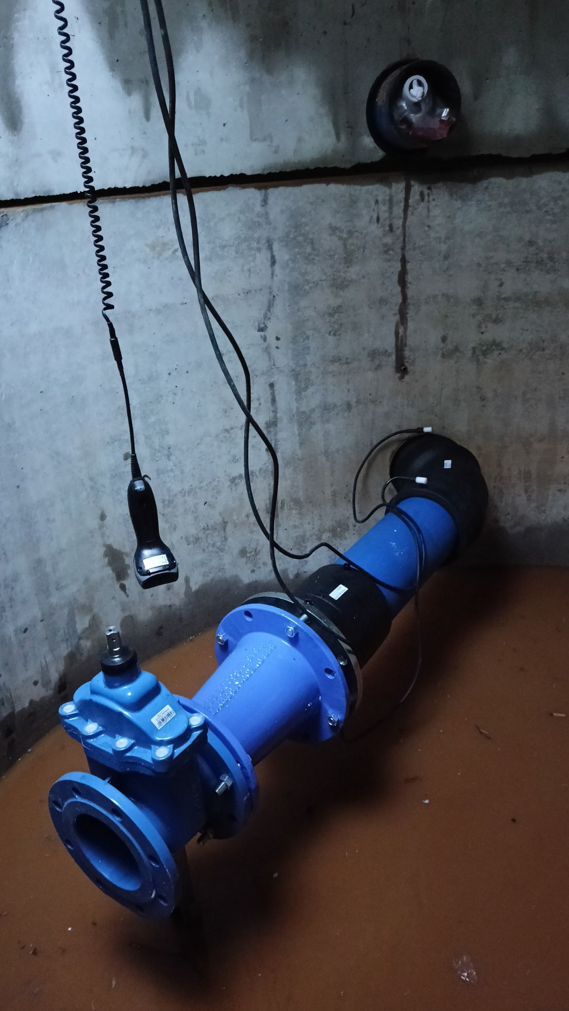 Niebieski zawór z podłączonymi przewodami w betonowym pomieszczeniu, prawdopodobnie studzience. Widoczny czujnik pomiarowy i woda na podłodze.