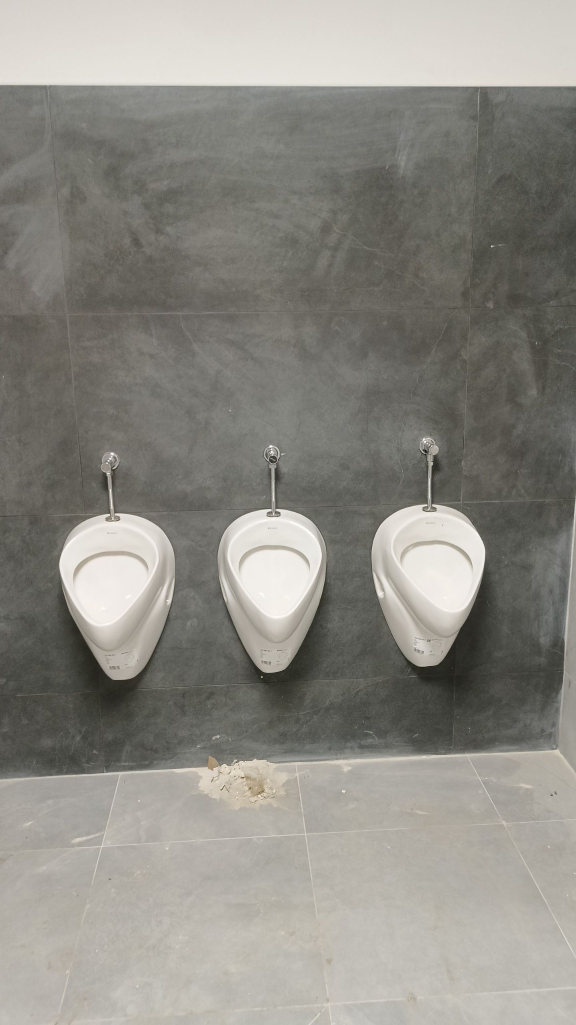 Trzy białe pisuary zamontowane na szarej, teksturowanej ścianie w rzędzie. Na podłodze widoczne ślady po montażu. Minimalistyczny design łazienki publicznej.