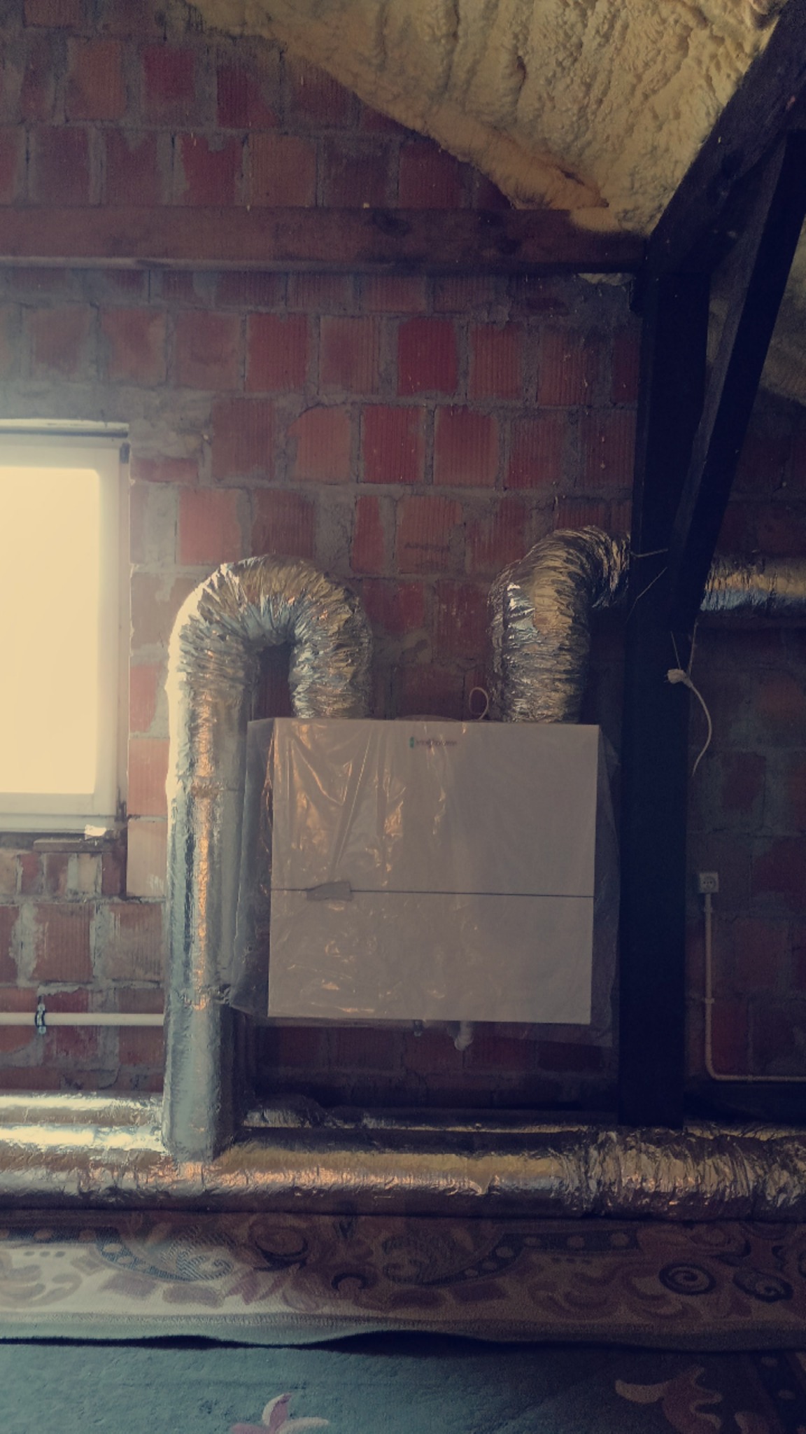 Rekuperator z aluminiowymi przewodami wentylacyjnymi w surowym wnętrzu poddasza z ceglaną ścianą i drewnianą konstrukcją dachu.