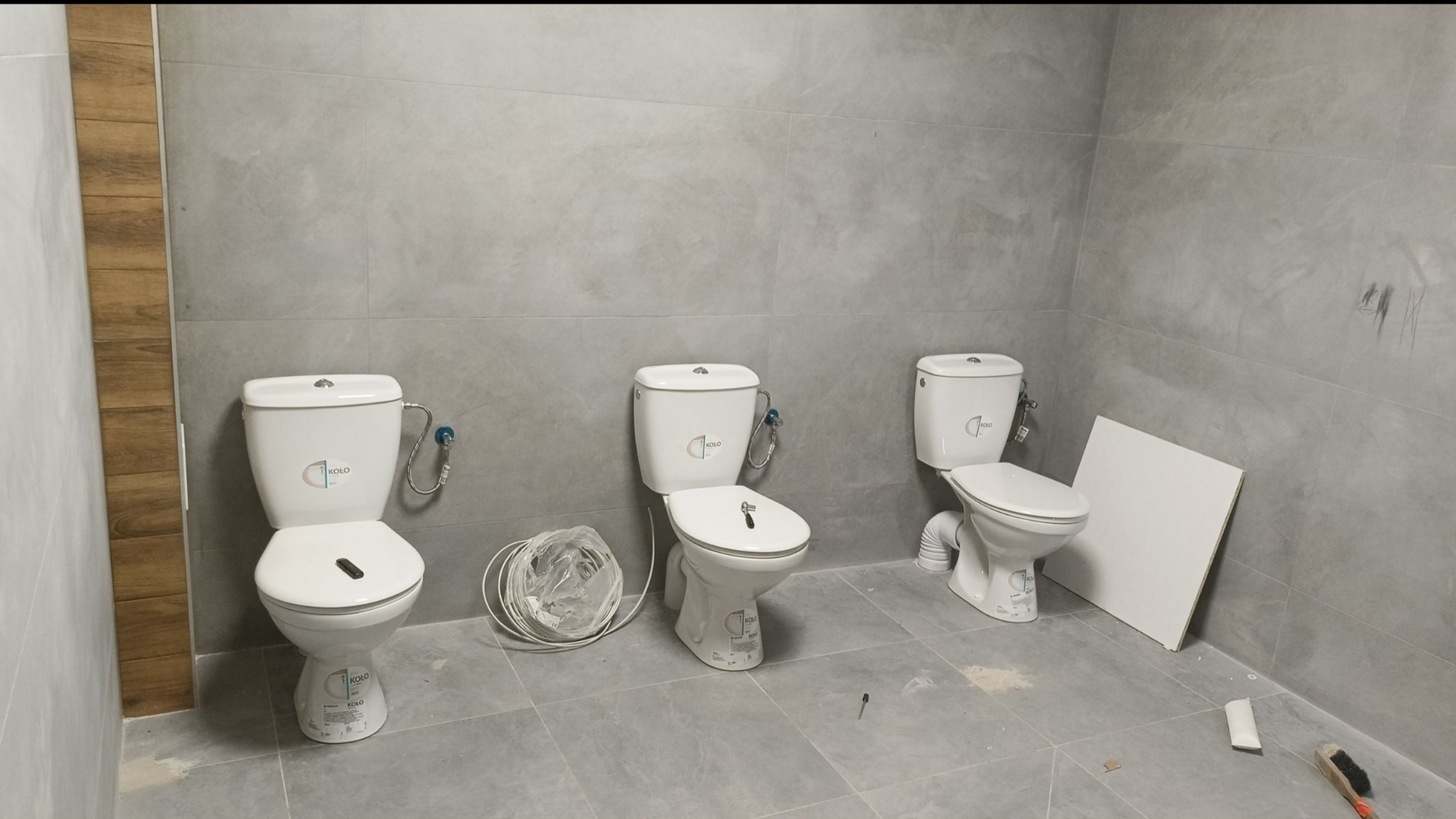 Trzy białe, częściowo zamontowane toalety Kolo w surowym wnętrzu łazienki z szarymi płytkami i drewnianym akcentem. Widoczne elementy instalacyjne i narzędzia.