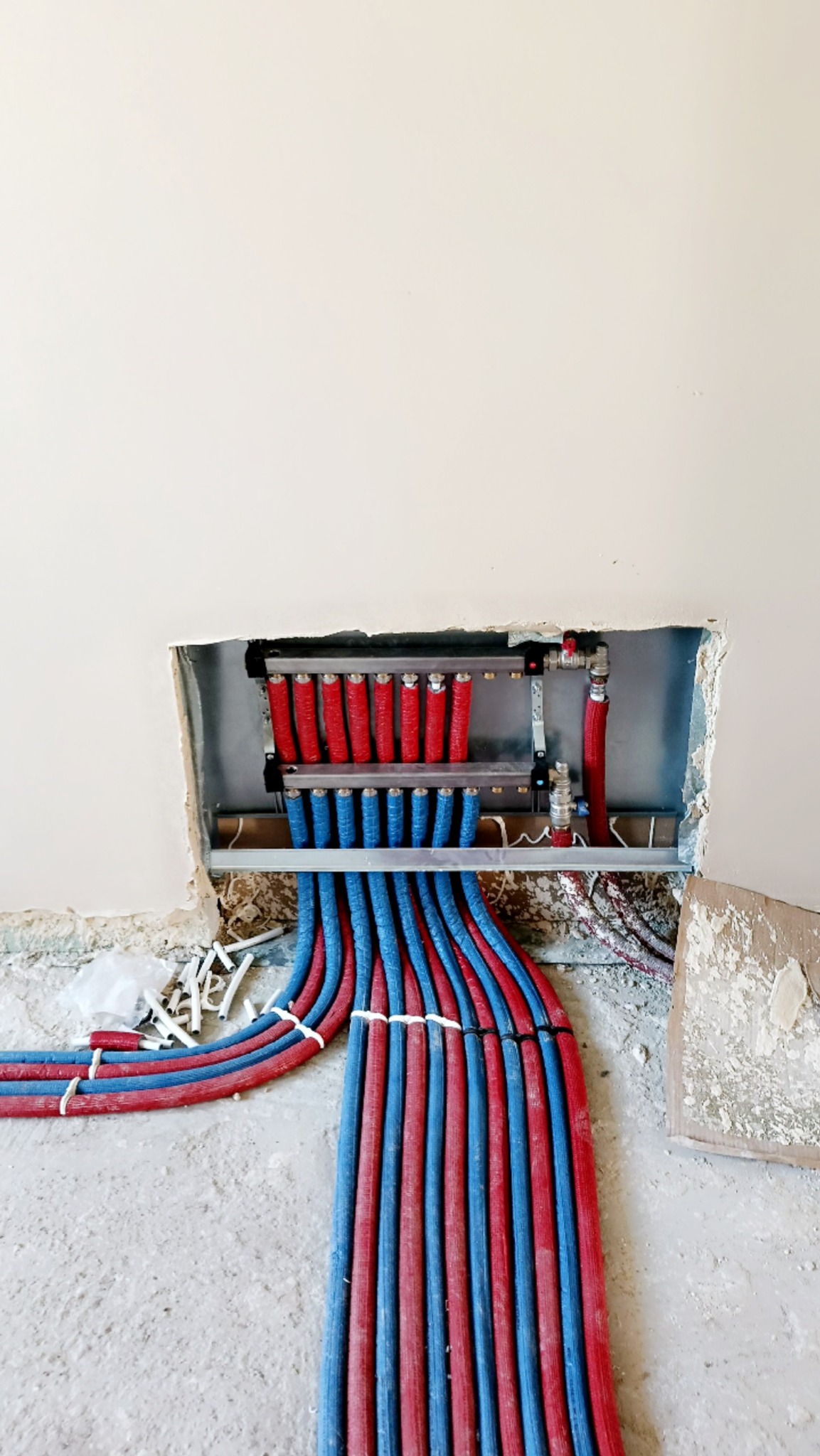 Rozdzielacz ogrzewania podłogowego z rurami w kolorze czerwonym i niebieskim, zamontowany w ścianie. Widoczne elementy instalacji i surowy stan podłoża.