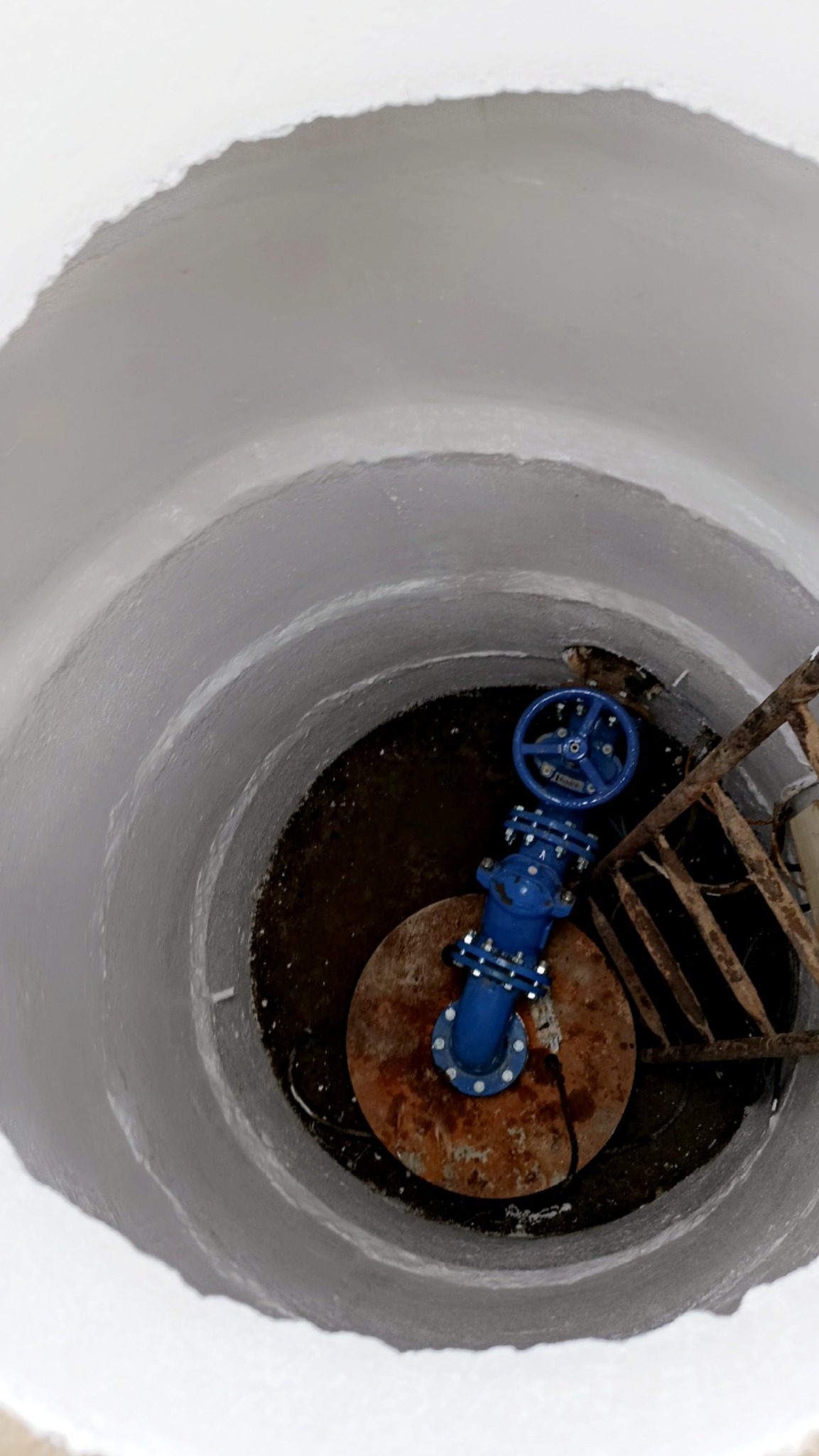 Niebieski zawór w studni z metalową drabinką, widok z góry. Białe, betonowe ściany studni kontrastują z rdzawą pokrywą i ciemnym dnem. Ujęcie z góry.