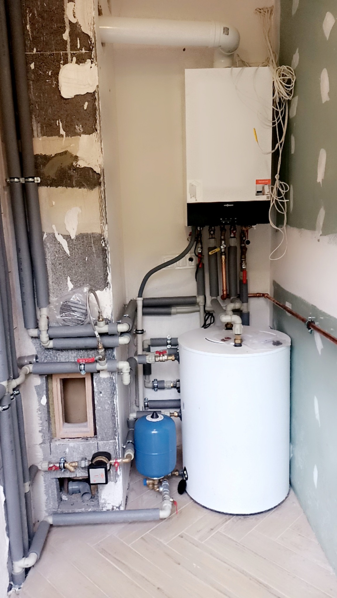 Biały bojler i piec Viessmann z szarymi rurami w pomieszczeniu w trakcie remontu. Widoczne niedokończone ściany i podłoga z jasnych paneli.