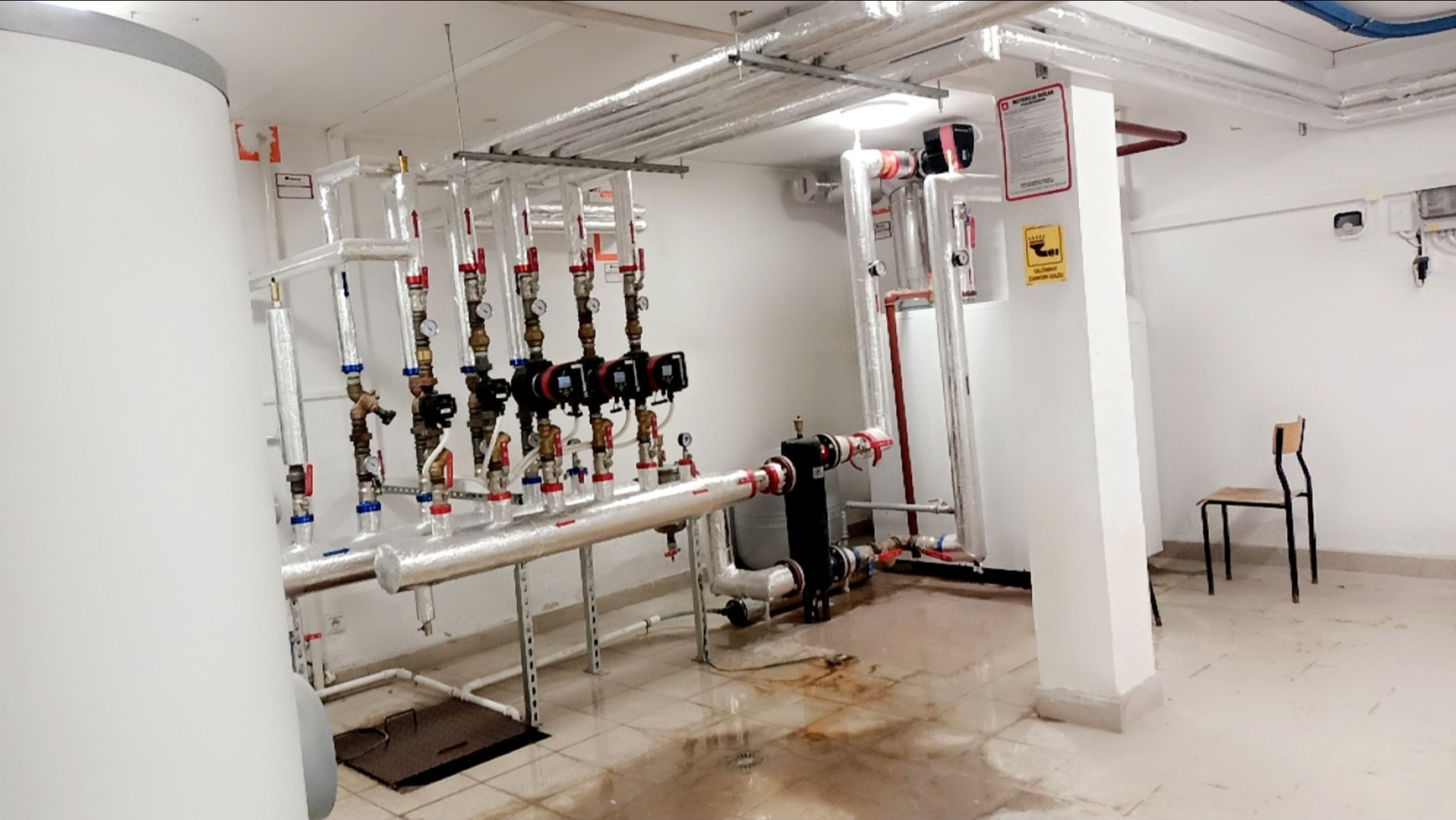 Złożona instalacja grzewcza z rurami w izolacji, zaworami i pompami obiegowymi w kotłowni. Widoczne elementy mocujące i tabliczki informacyjne. Na podłodze widoczne ślady wilgoci.
