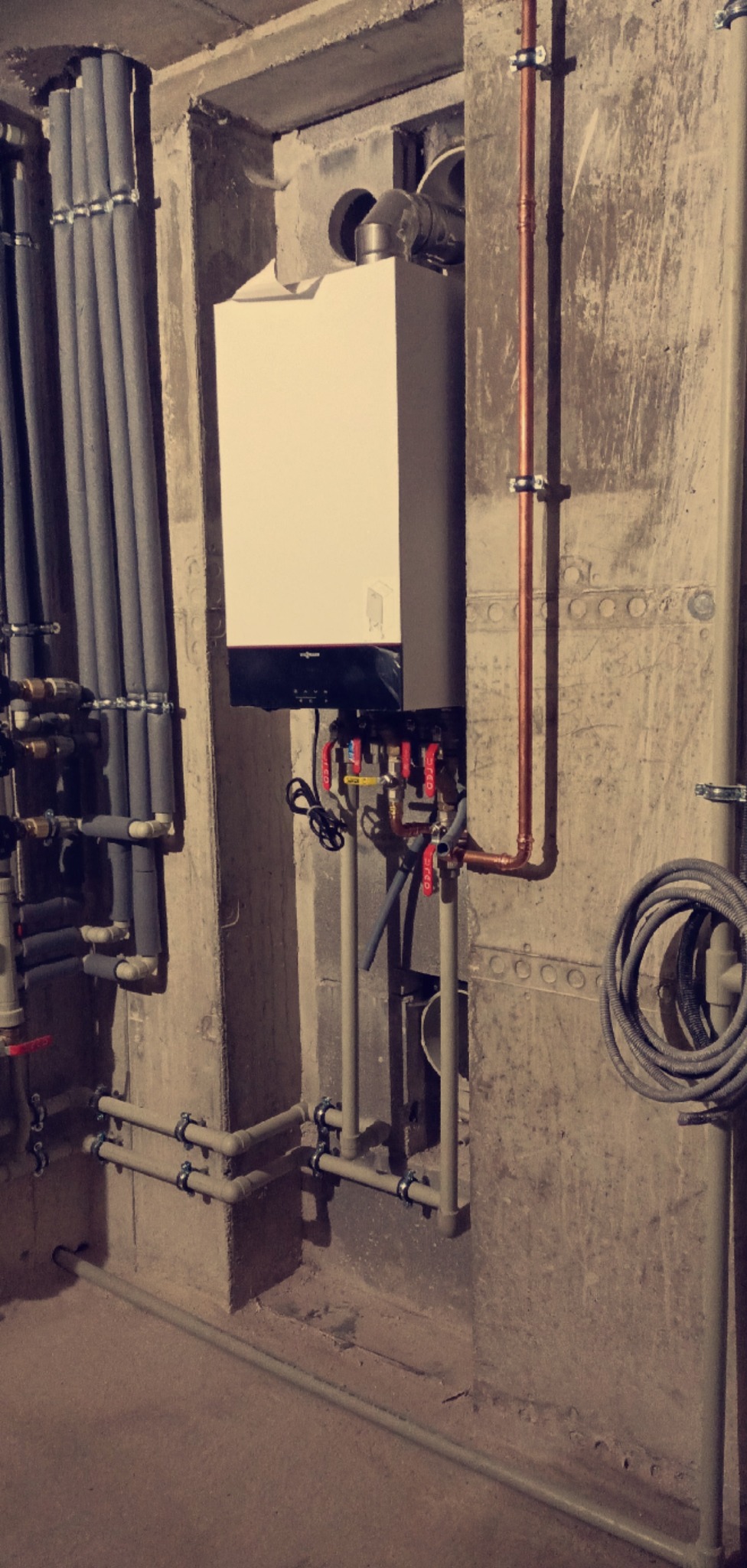 Biały piec grzewczy Viessmann zamontowany na betonowej ścianie, z widocznymi rurami miedzianymi i szarymi, zaworami oraz elementami instalacji w surowym pomieszczeniu.