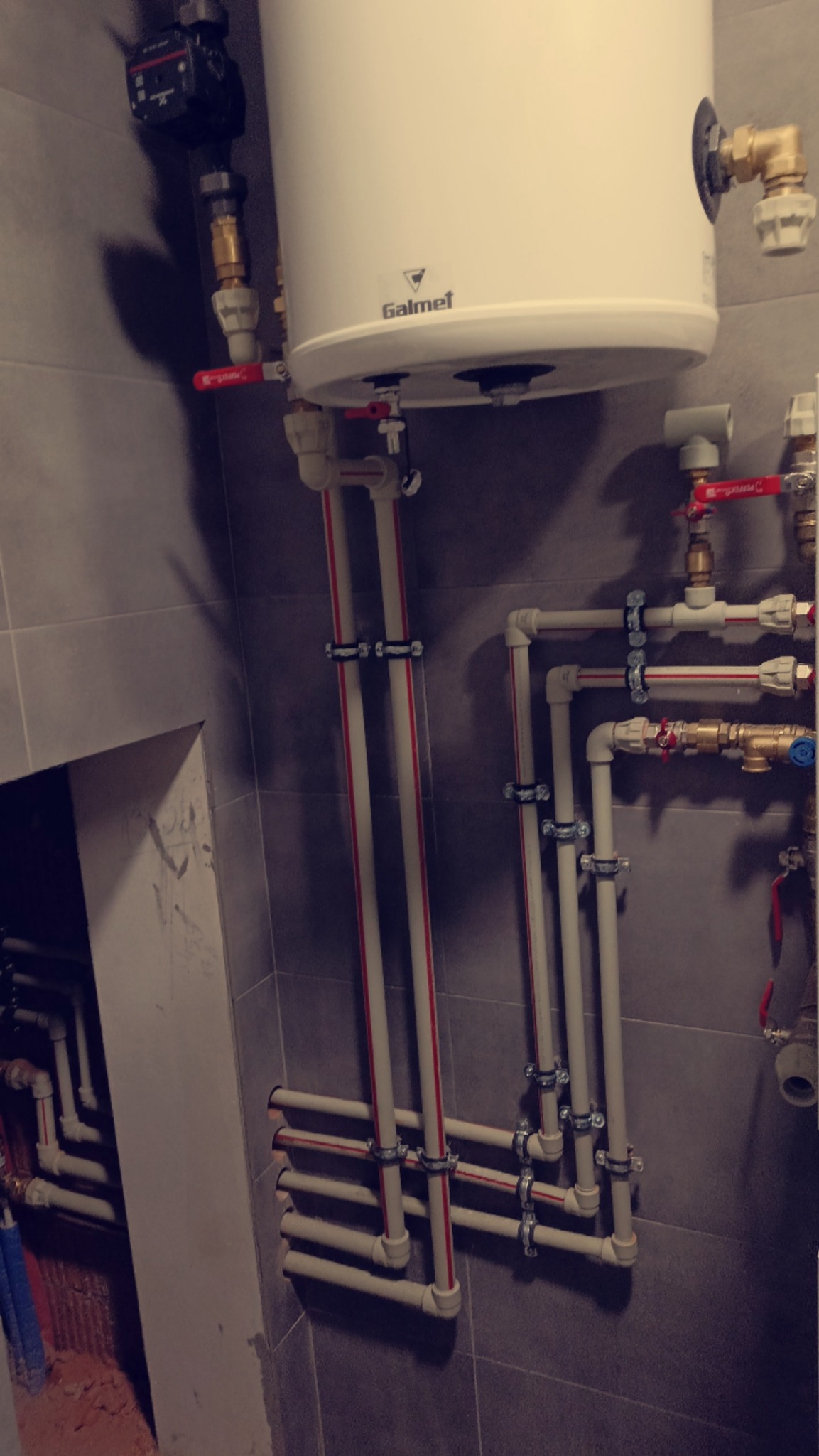 Biały bojler Galmet z podłączeniami rur PCV i zaworami na szarej, kafelkowanej ścianie. Widoczne elementy instalacji hydraulicznej w pomieszczeniu.