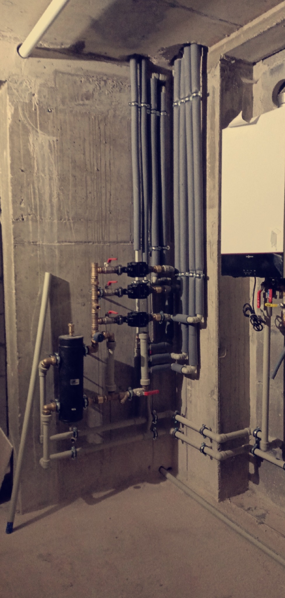 Złożona instalacja grzewcza z rurami w izolacji, zaworami i kotłem w surowym wnętrzu. Widoczne elementy hydrauliczne i rury prowadzone wzdłuż betonowej ściany.