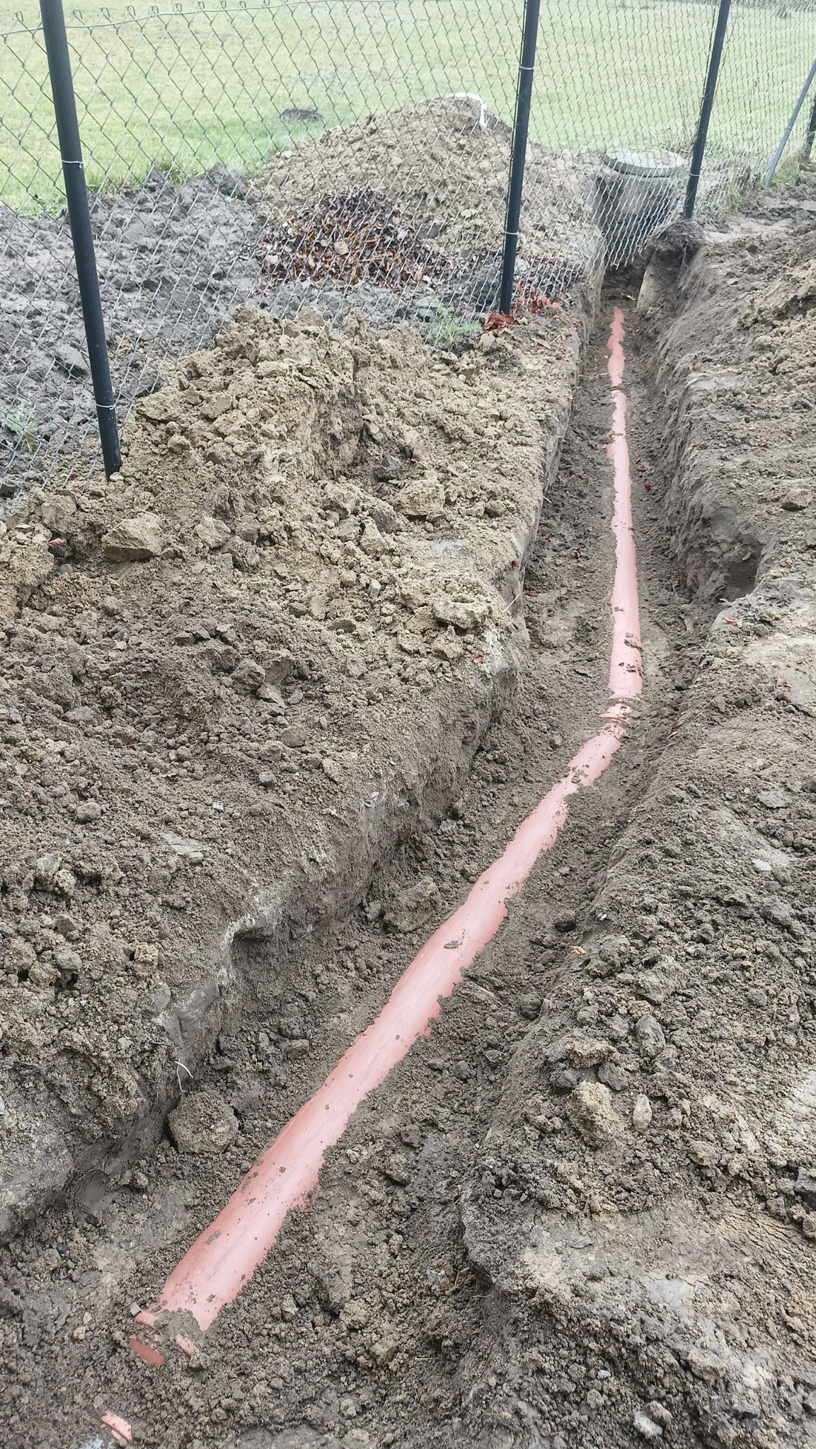 Rów z ułożoną rurą z tworzywa sztucznego, widoczny fragment ogrodzenia z siatki. Wykop w ziemi, przygotowanie do zakopania instalacji.