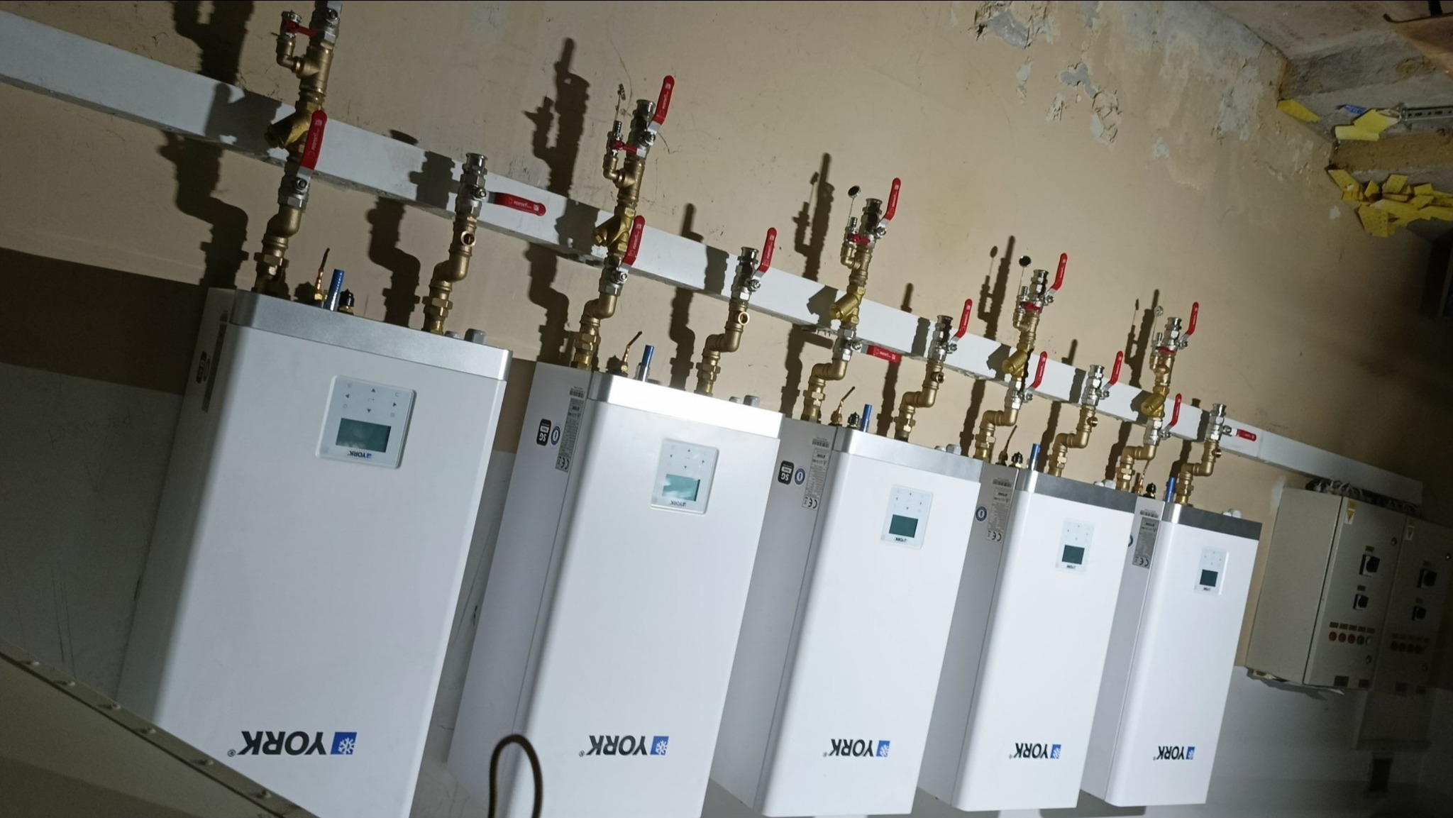 Rząd białych, wiszących kotłów grzewczych marki York z mosiężnymi rurami i zaworami, zamontowanych na ścianie z widocznymi śladami użytkowania.