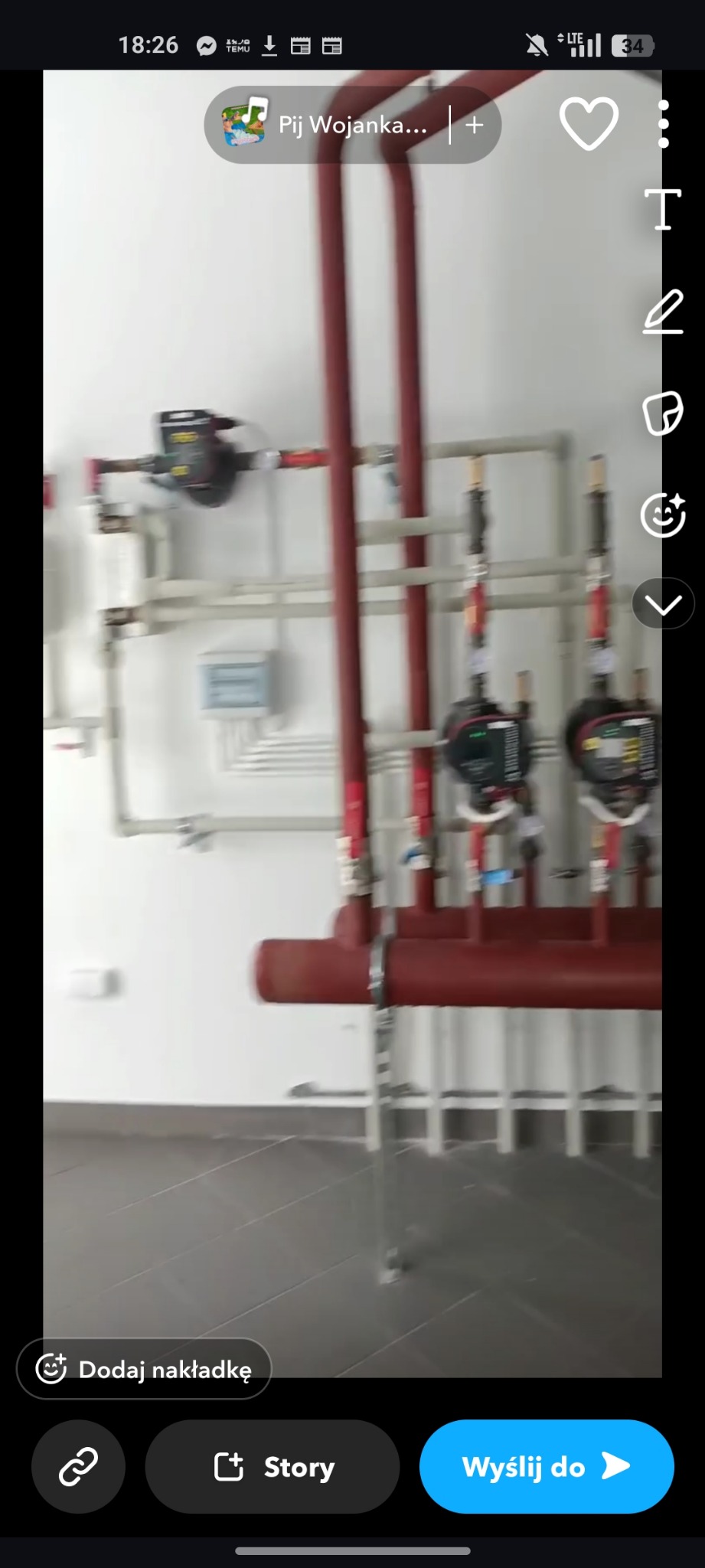 Złożona instalacja grzewcza z czerwonymi rurami i czarnymi zaworami na białej ścianie. Widoczne liczne rury i elementy instalacji, szara podłoga z płytek.