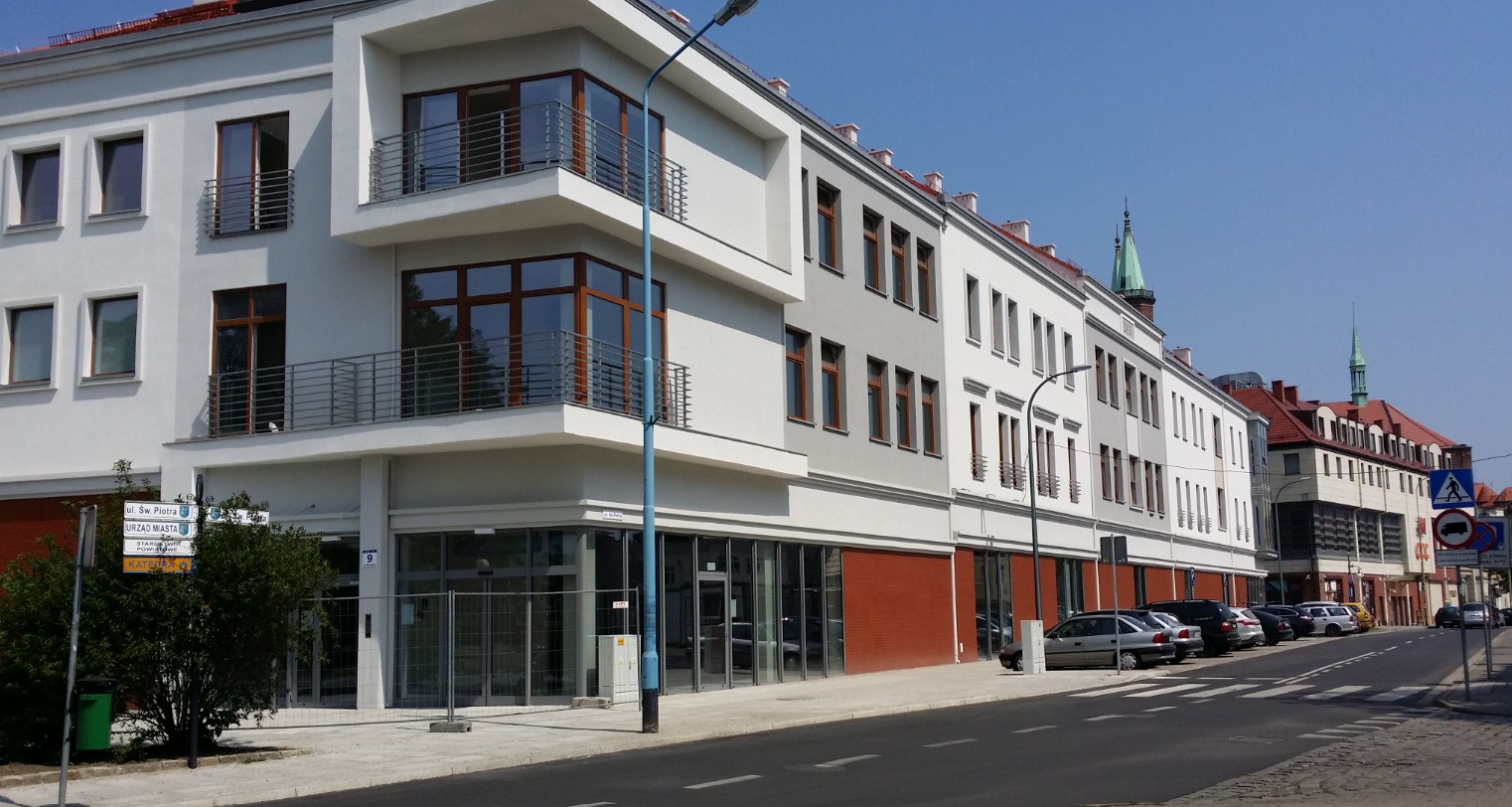 Nowoczesny budynek biurowy z balkonami i dużymi oknami, widok z ulicy w słoneczny dzień. Czerwona cegła na parterze, szara elewacja na wyższych kondygnacjach.