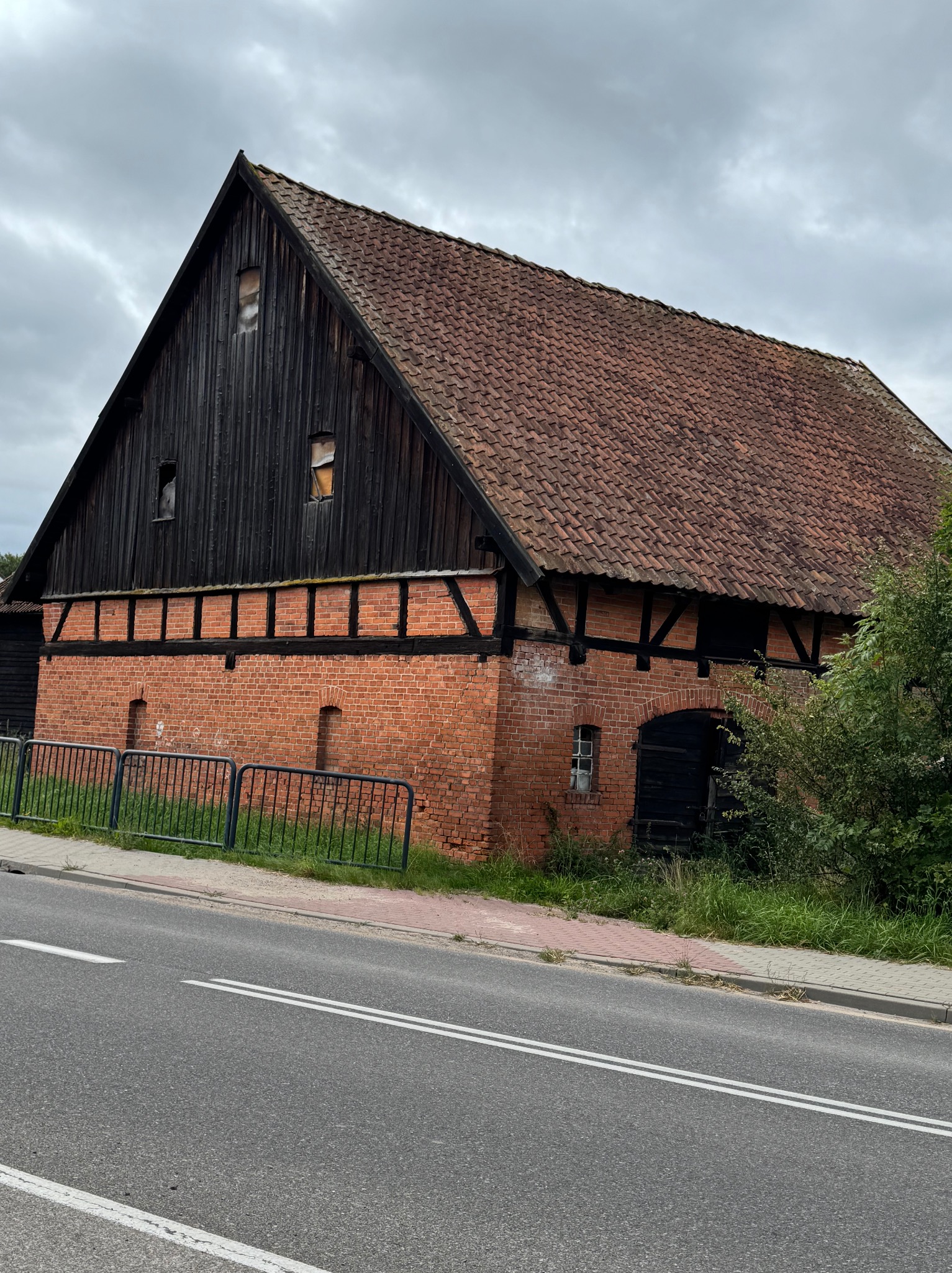 Stary, ceglano-drewniany budynek gospodarczy z czerwoną dachówką, widoczny z poziomu ulicy. Fragment metalowego ogrodzenia i zarośnięte otoczenie dodają rustykalnego charakteru.