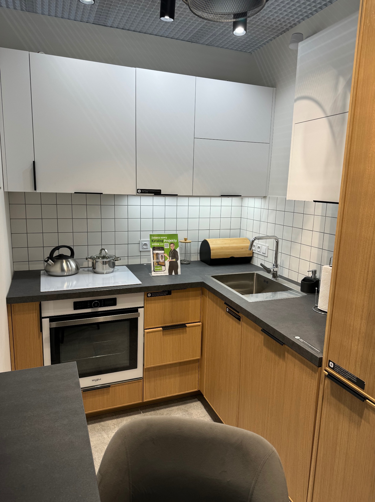 Nowoczesna, jasna kuchnia z drewnianymi szafkami i białymi górnymi frontami. Na blacie czajnik, garnek i akcesoria kuchenne. Minimalistyczny design z płytkami w tle.