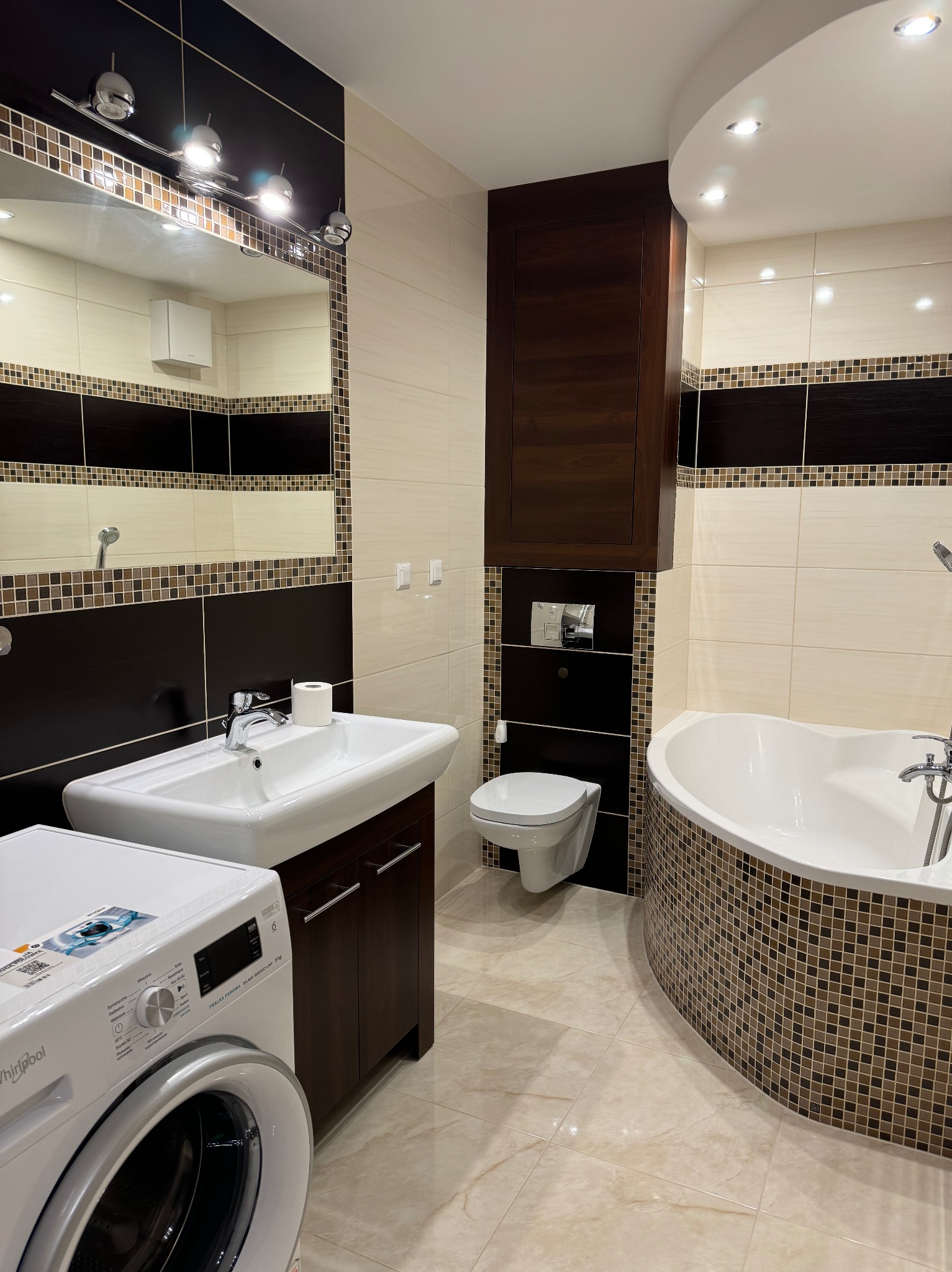 Nowoczesna łazienka z beżowo-brązowymi płytkami mozaikowymi, białą armaturą (umywalka, toaleta, wanna) i pralką Whirlpool. Ciemne, drewniane szafki i lustro z oświetleniem.