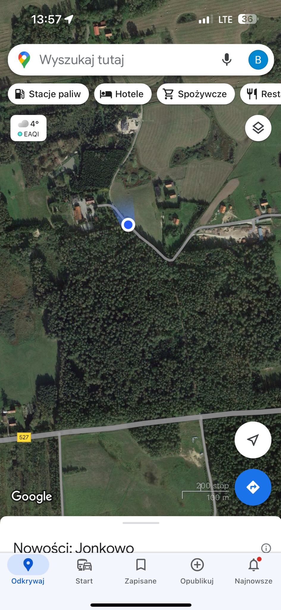 Widok z lotu ptaka na teren zalesiony i zabudowania, prezentowany na ekranie smartfona z aplikacją mapową. Widoczne elementy interfejsu aplikacji.