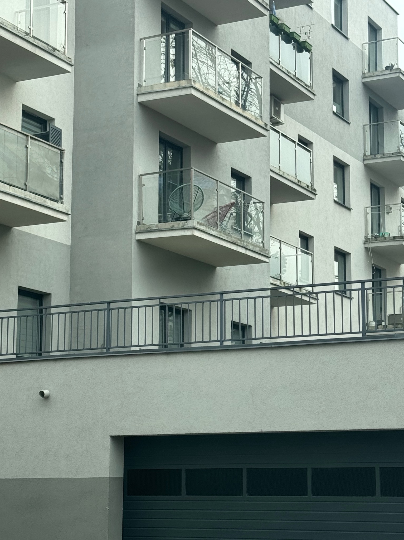 Nowoczesny budynek mieszkalny z balkonami o szklanych balustradach. Na jednym z balkonów widoczne krzesła. Ujęcie z dołu, widoczny fragment garażu.