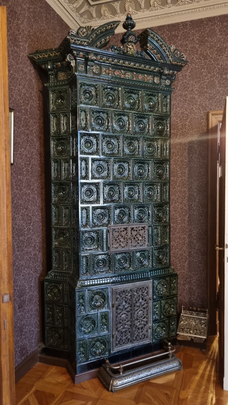 Stylowy, wysoki piec kaflowy w odcieniach zieleni i złota, z bogatymi zdobieniami, na tle ściany w brązowe wzory. Widoczny fragment drewnianej podłogi i drzwi.