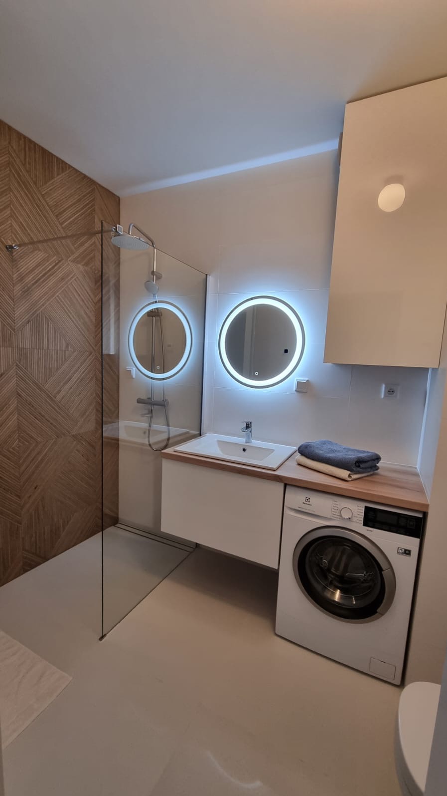 Nowoczesna łazienka z kabiną prysznicową, okrągłymi lustrami z podświetleniem LED i pralką. Drewniana ściana z geometrycznym wzorem dodaje ciepła.