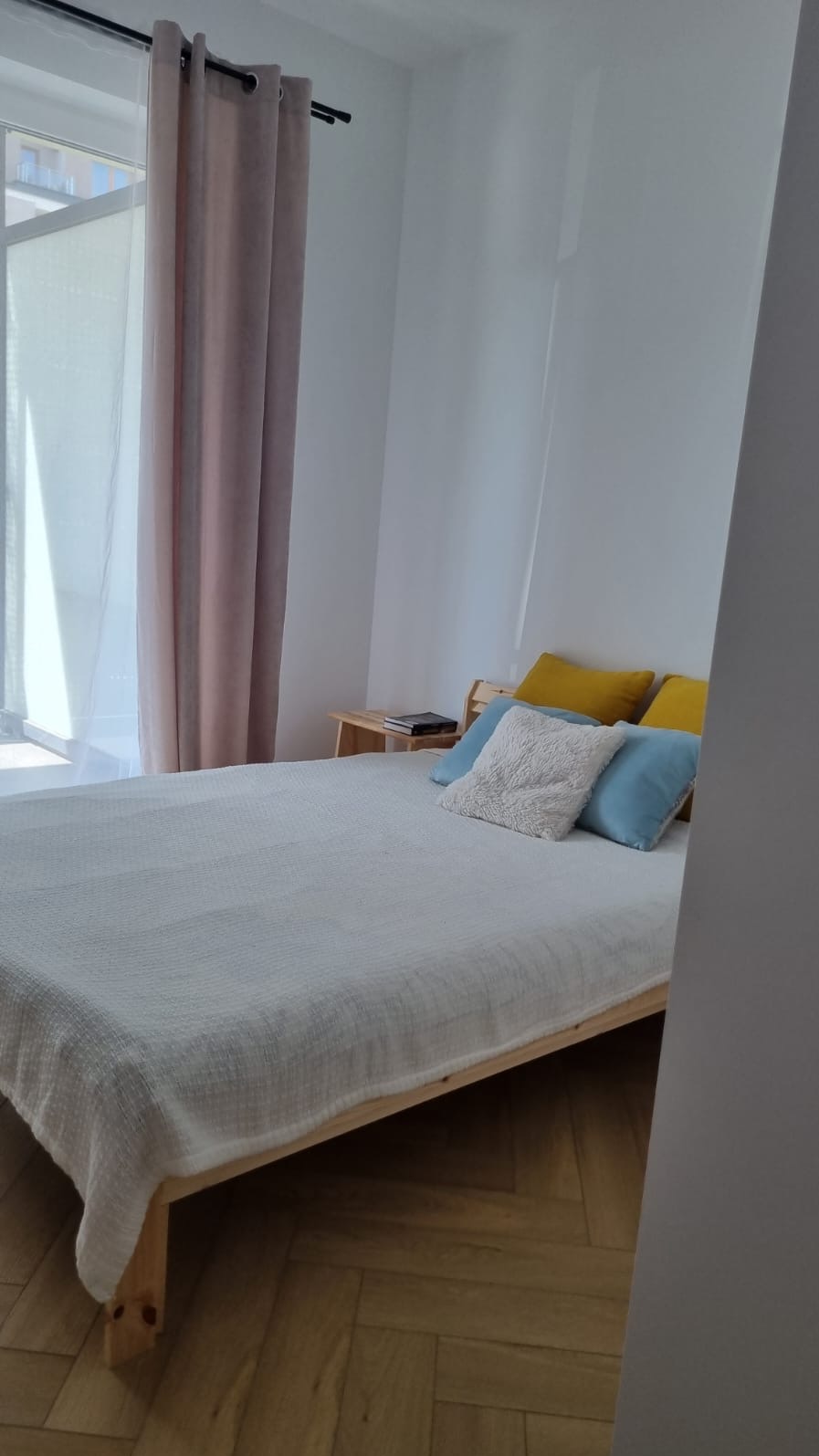 Jasna sypialnia z drewnianym łóżkiem, białym pledem i kolorowymi poduszkami. Parkiet w jodełkę, okno z zasłonami. Minimalistyczny styl wnętrza.