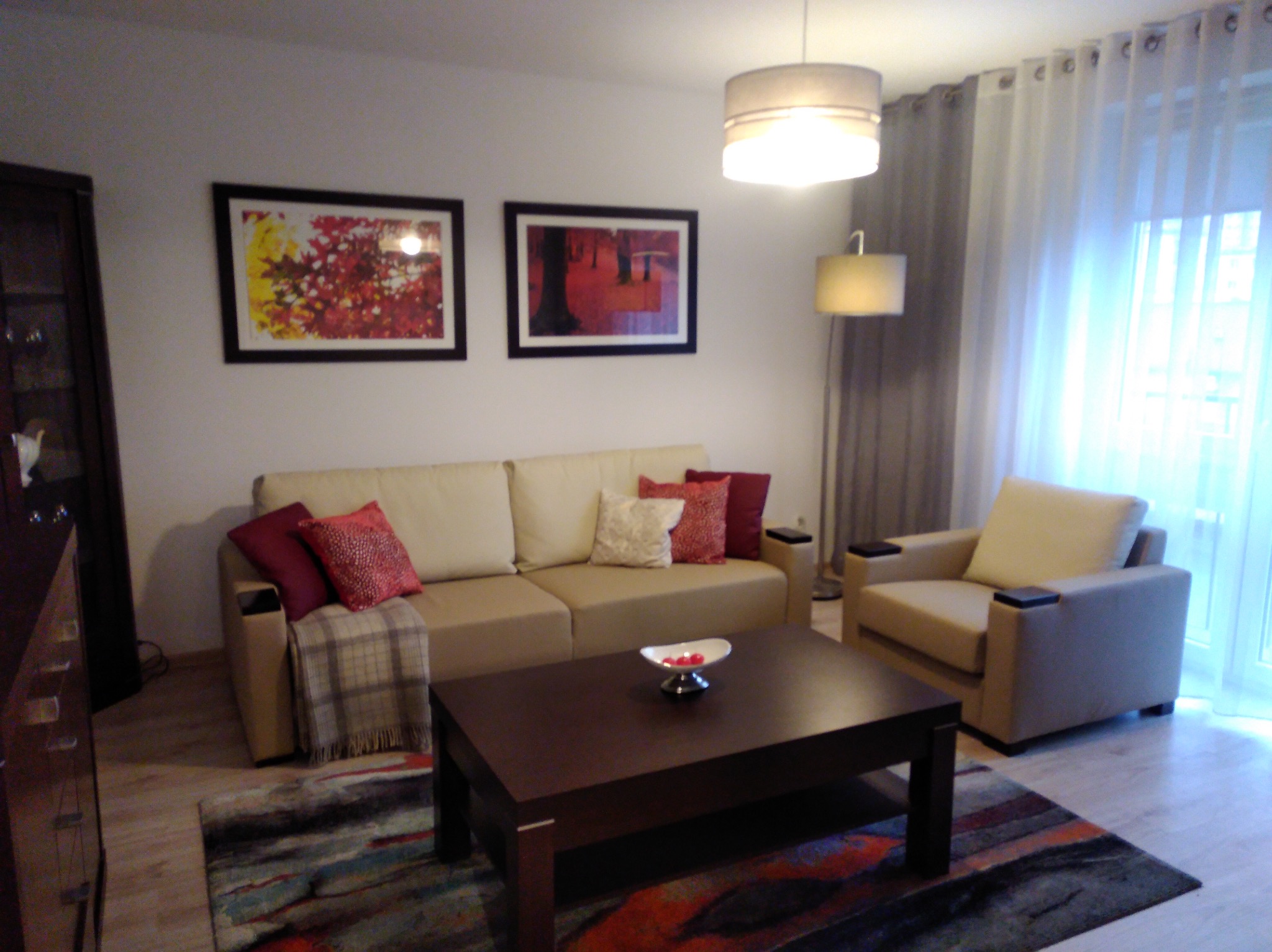 Jasny salon z beżową sofą, fotel, ciemny stolik kawowy i kolorowy dywan. Dwa obrazy na ścianie i lampa podłogowa. Okno z zasłonami w tle.