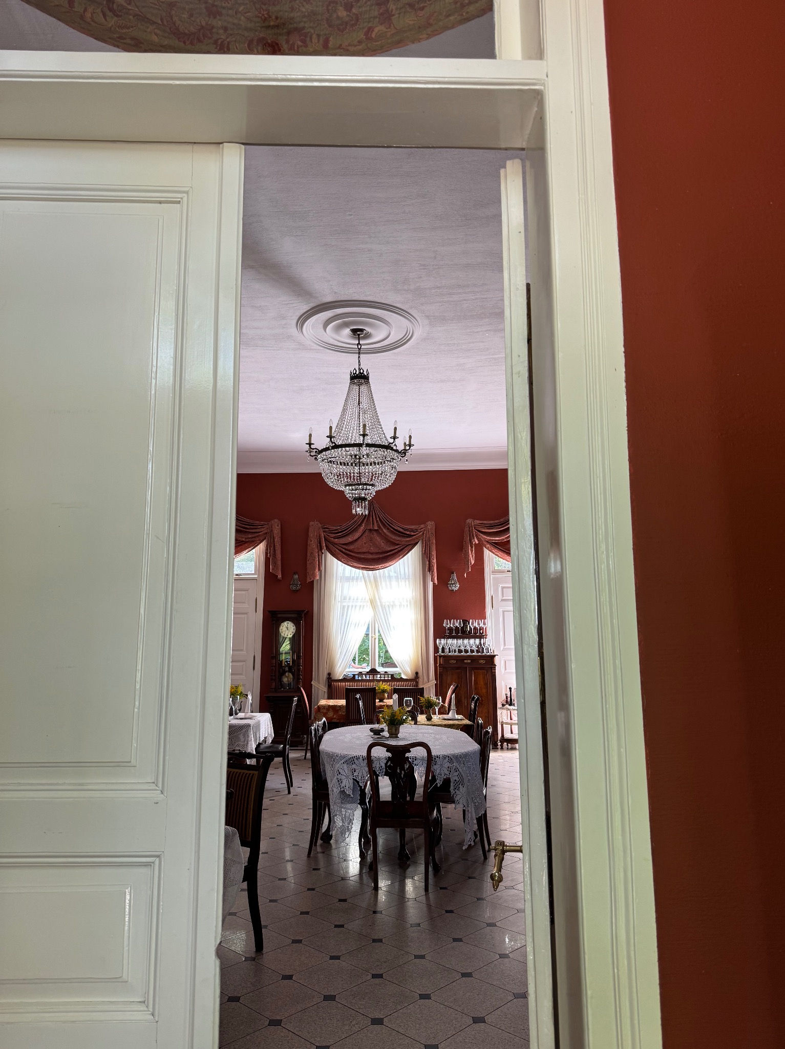 Elegancka jadalnia z kryształowym żyrandolem widziana przez otwarte drzwi. Czerwone ściany, stół z koronkowym obrusem, antyczne meble i zegar.