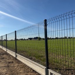 GRAFIT BARTOSZ DZIUBAŃSKI - Nowoczesne ogrodzenie panelowe z podmurówką betonową na tle zielonego pola i błękitnego nieba. Widoczne słupy i siatka w kolorze grafitowym. Słoneczny dzień.