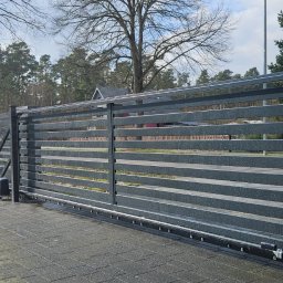 GRAFIT BARTOSZ DZIUBAŃSKI - Nowoczesna, przesuwna brama wjazdowa z poziomymi panelami, zintegrowana ze słupkiem z szarego kamienia. Widoczny automat do bramy i fragment ogrodzenia panelowego.
