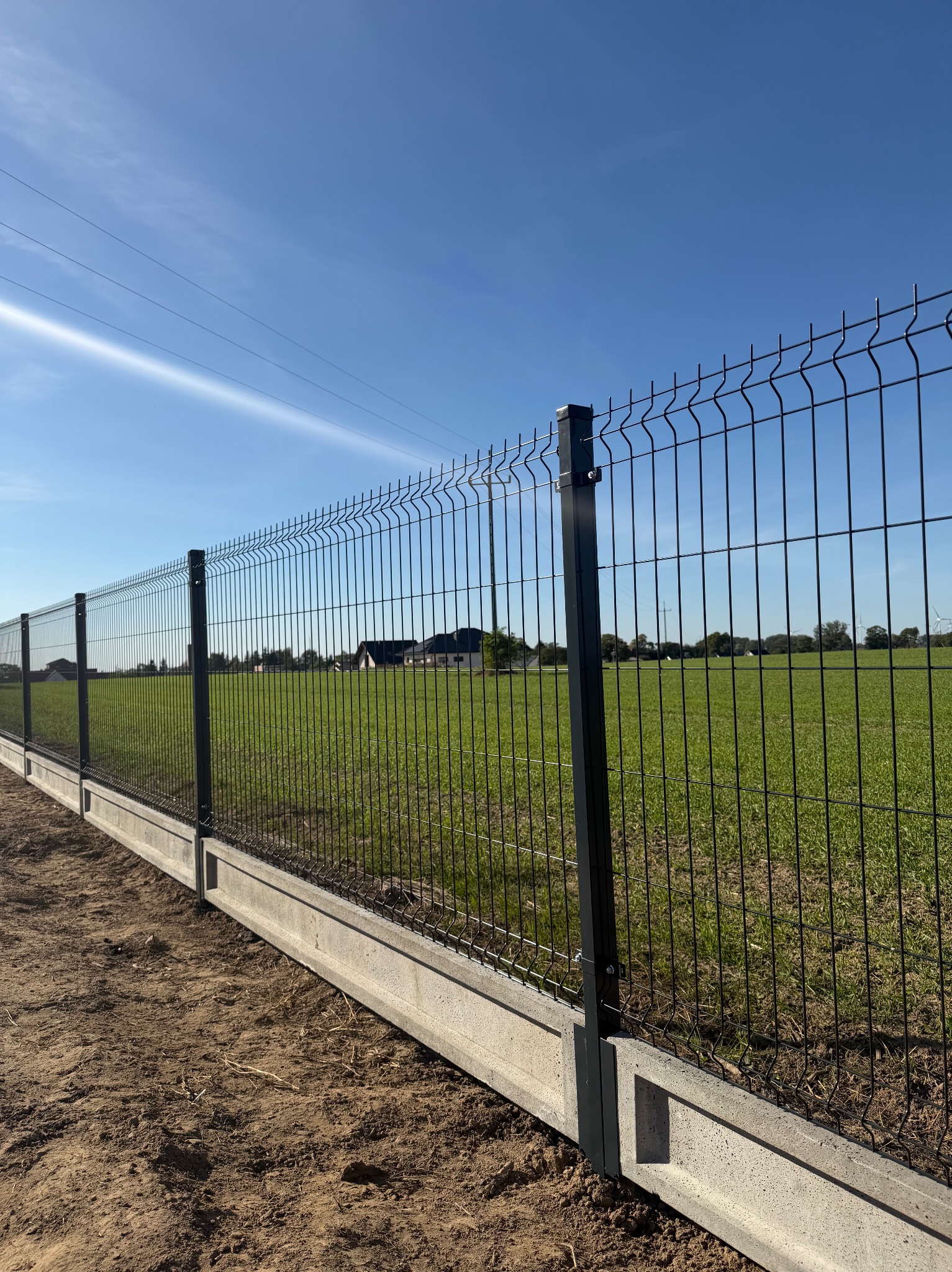 Nowoczesne ogrodzenie panelowe z podmurówką betonową na tle zielonego pola i błękitnego nieba. Widoczne słupy i siatka w kolorze grafitowym. Słoneczny dzień.