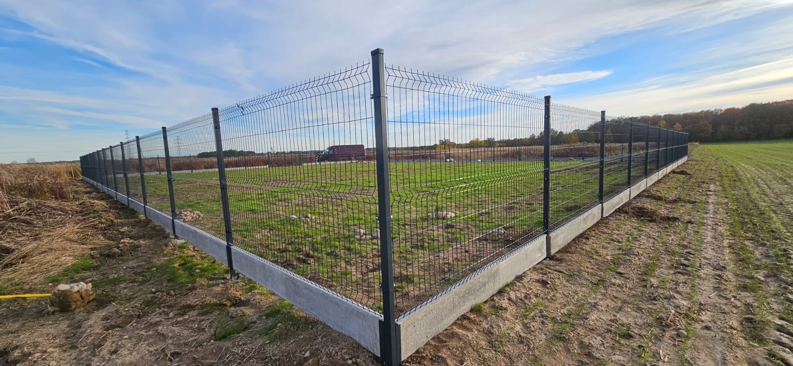 Kompletne ogrodzenie panelowe z podmurówką betonową, otaczające zielony teren. Widoczne słupki i siatka w kolorze grafitowym, błękitne niebo w tle.