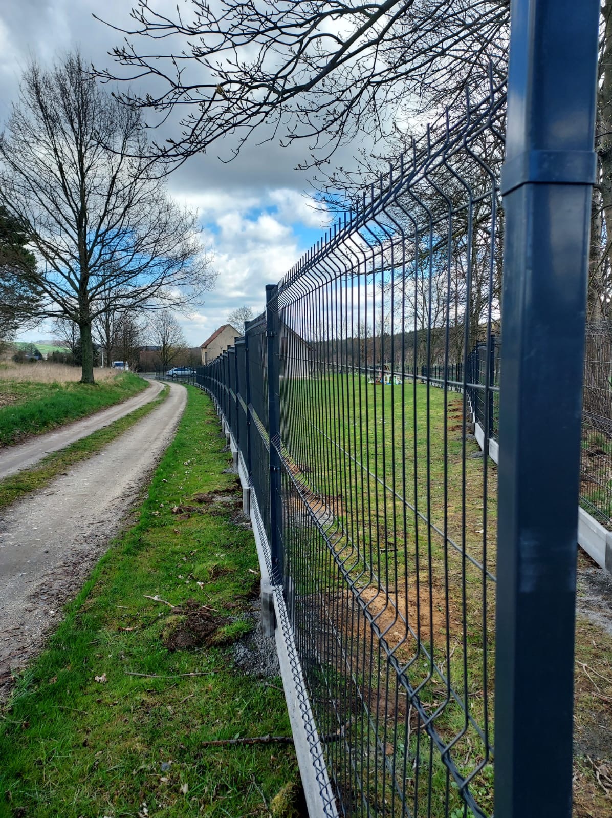 Długi, ciemny panel ogrodzeniowy z podmurówką betonową, biegnący wzdłuż polnej drogi. Widok na zieloną trawę i drzewa w tle, pochmurne niebo.