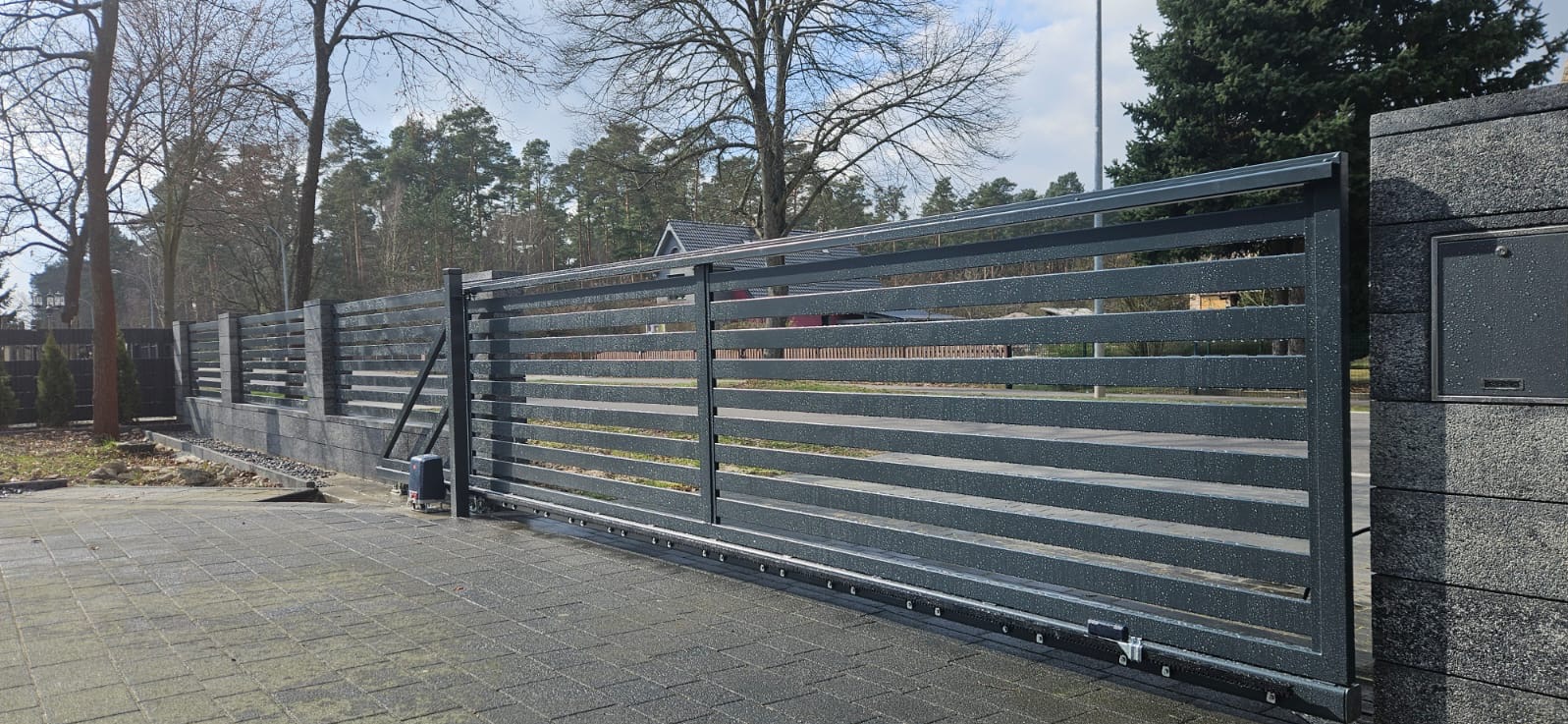 Nowoczesna, przesuwna brama wjazdowa z poziomymi panelami, zintegrowana ze słupkiem z szarego kamienia. Widoczny automat do bramy i fragment ogrodzenia panelowego.