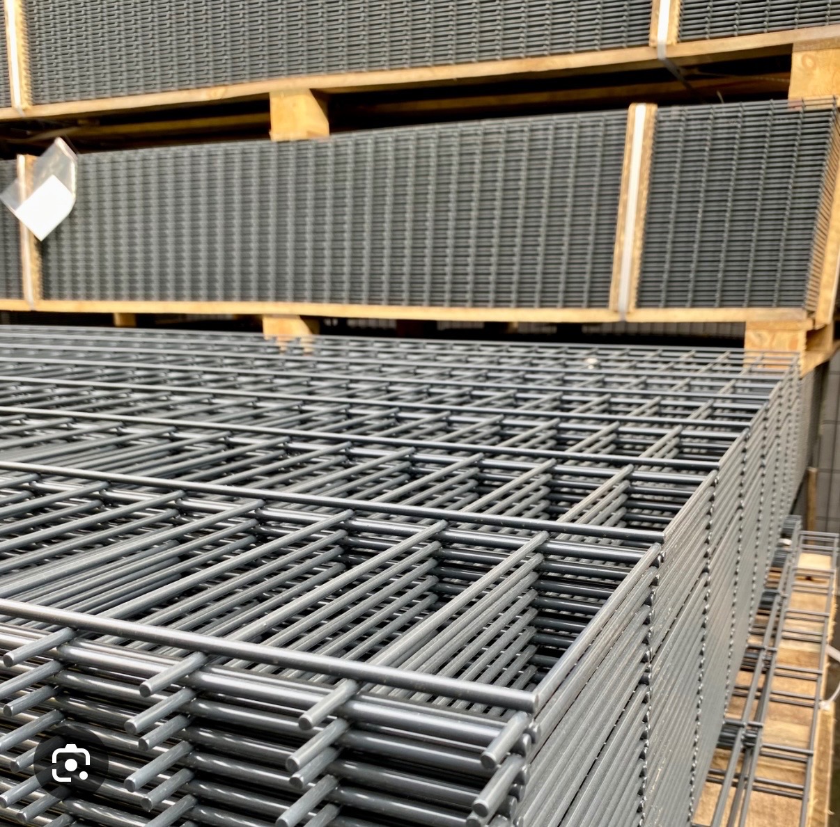 Stos paneli zgrzewanych z drutu na palecie. Szare panele tworzą regularną siatkę. Widoczne spawy na łączeniach drutów. Magazynowe oświetlenie.