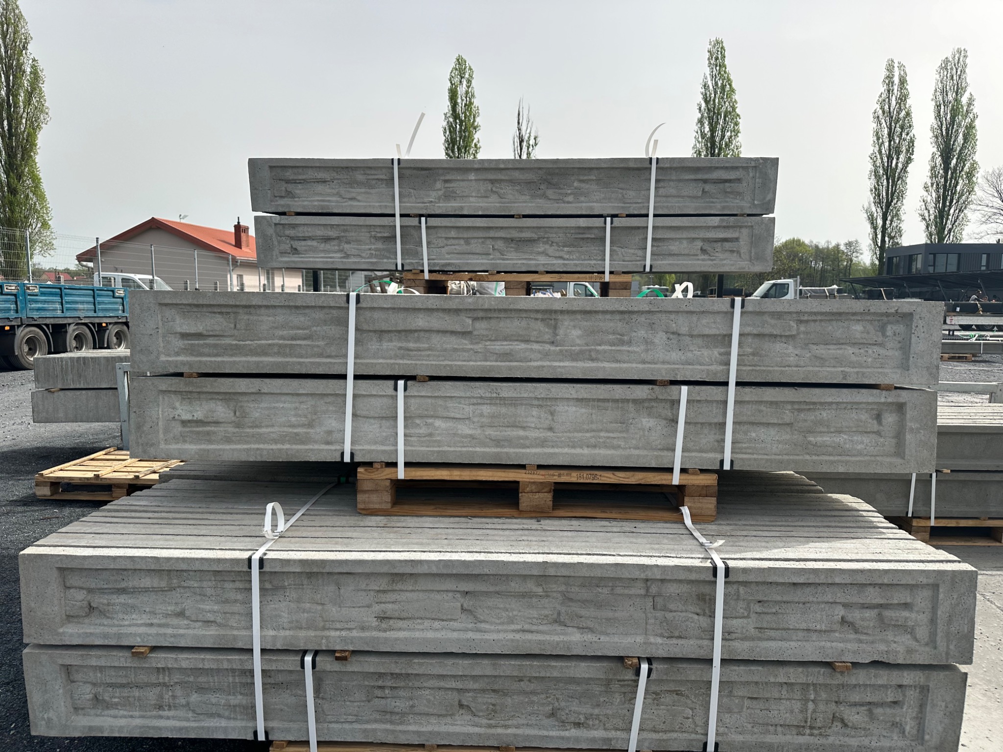 Ułożone na paletach betonowe elementy ogrodzeniowe z dekoracyjnym wzorem, zabezpieczone białymi taśmami. W tle budynki i drzewa.